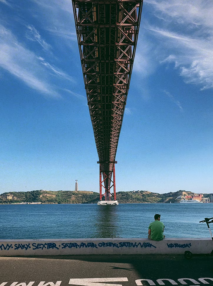 View under a massive red suspension bridge with a person sitting by the waterfront and the Christ the King statue visible across the river.