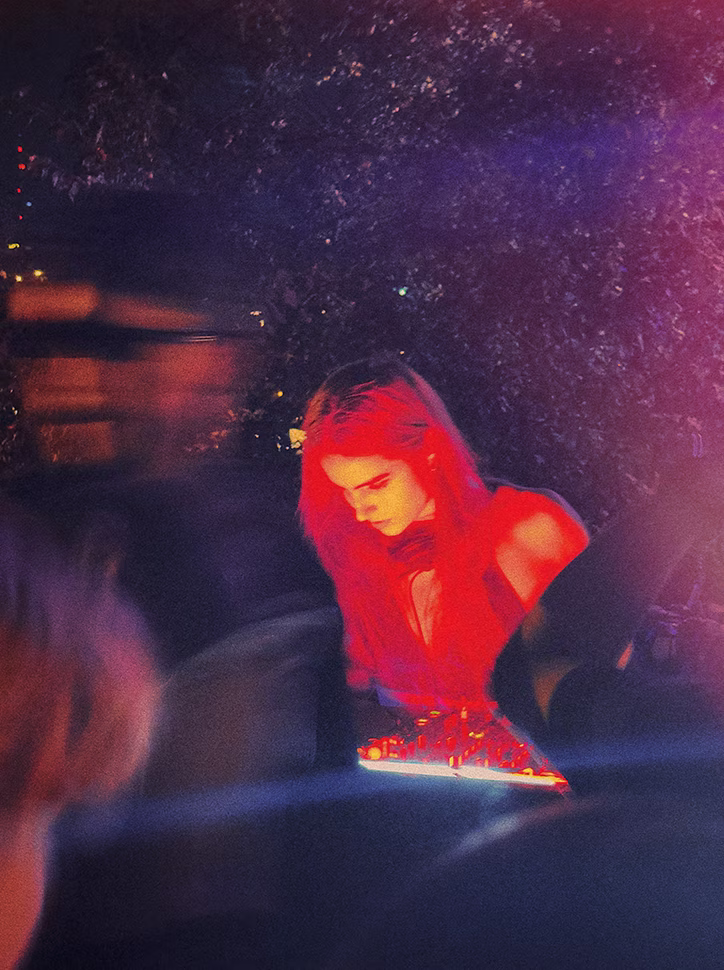Woman performing electronic music under red and purple lighting in a dark outdoor setting.