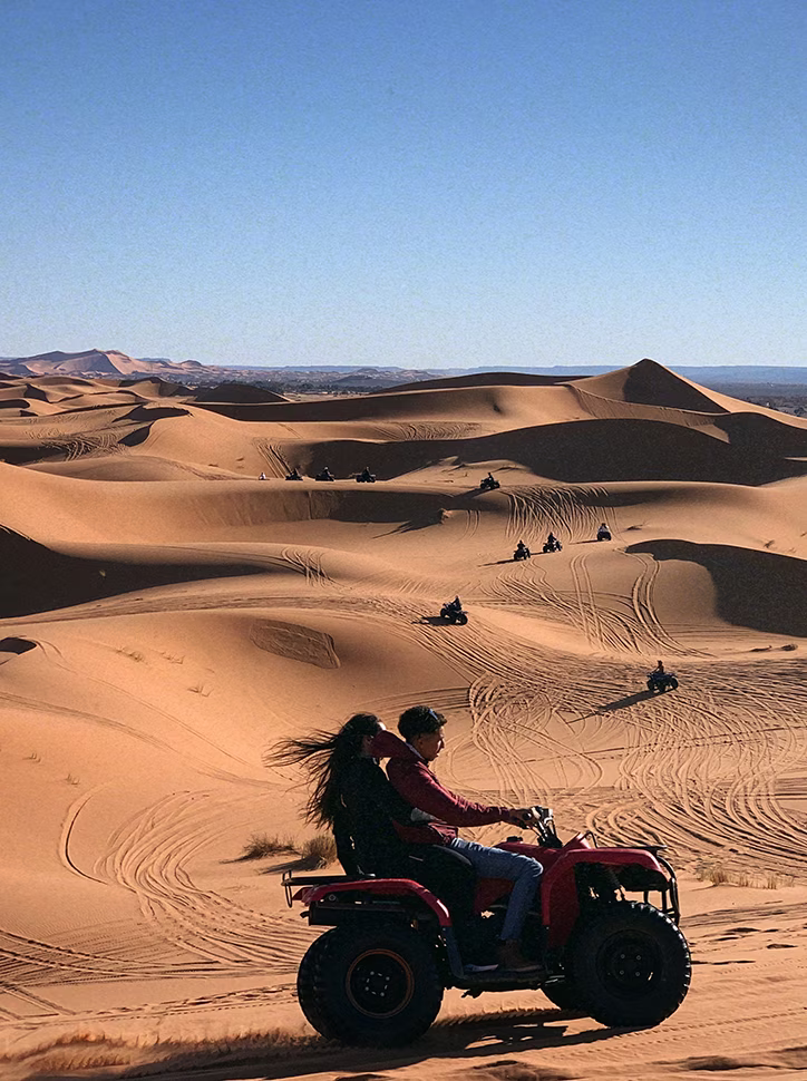 Couple riding an ATV through vast desert sand dunes with tire tracks and several other riders in the distance.