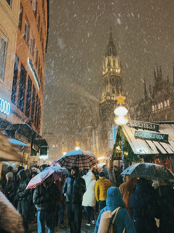 Crowd of people with umbrellas walking through a snowy Christmas market at night, with a tall illuminated Gothic-style tower in the background.