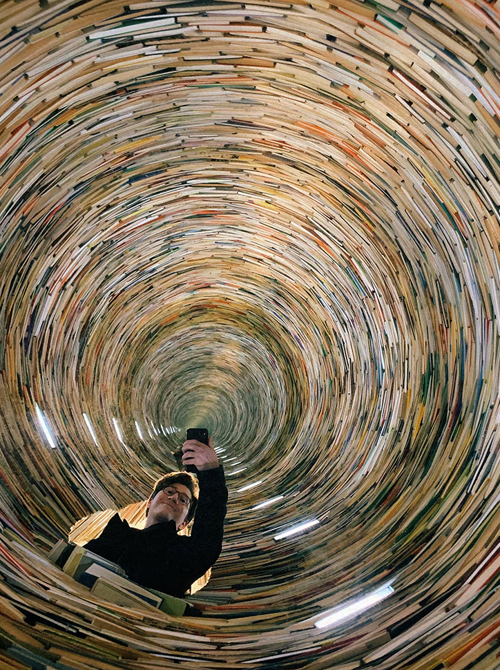 Roman Chekurov taking a selfie in a circular tunnel made of stacked books that spirals infinitely upward.