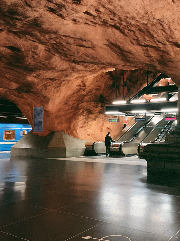 Underground metro station with orange cave-like walls and a person standing near the escalators beside a blue train.